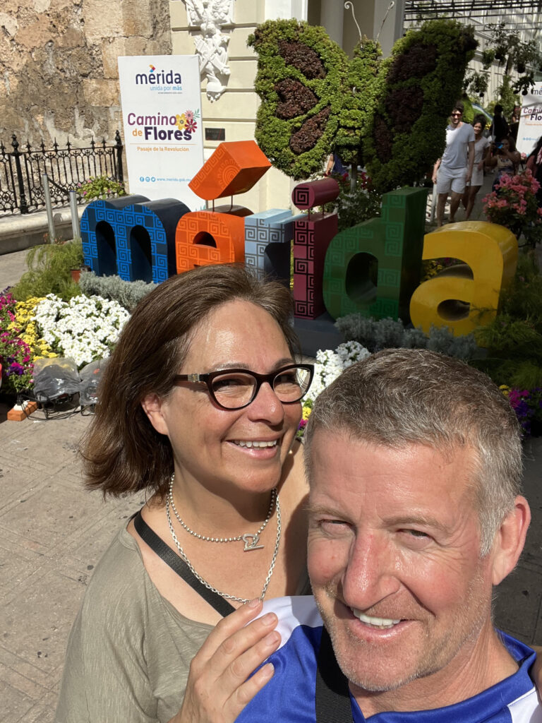 a woman and a man smiling in front of colourful Merida sign in Mexico