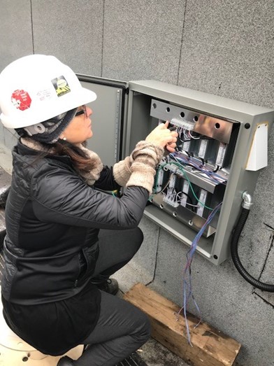 a women, wearing a hardhat, is crouched down working on an electrical panel
